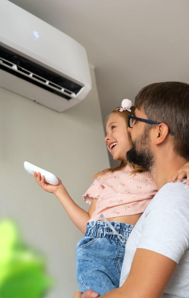 Père tenant sa fille souriante, elle règle la clim sur 16 avec la télécommande. Rires pour la fraîcheur estivale.