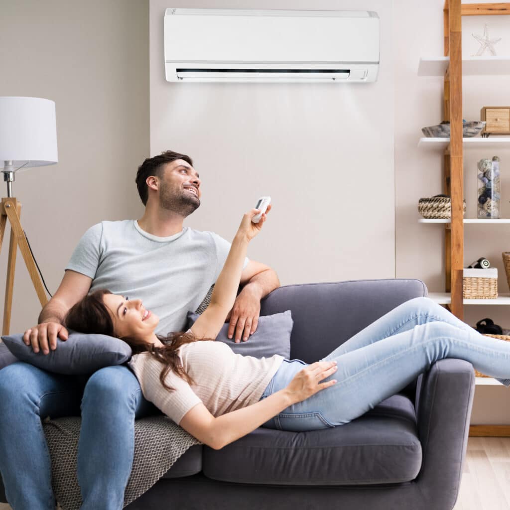 Couple souriant sur un canapé, la femme allongée sur l'homme qui règle un climatiseur mural avec une télécommande.