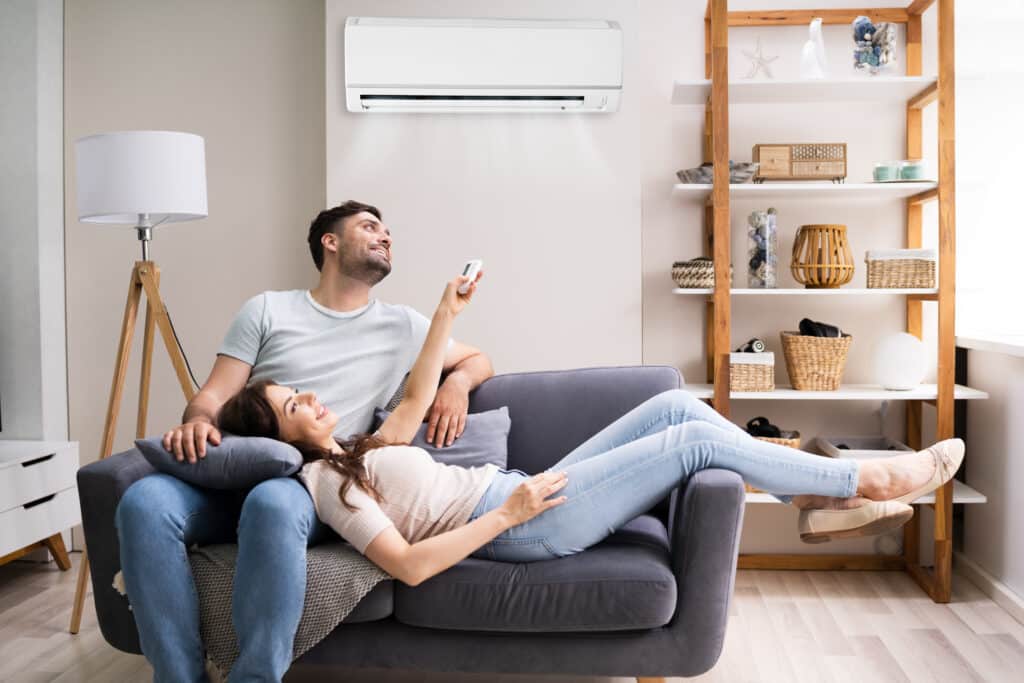 Couple souriant réglant le climatiseur avec une télécommande, la femme allongée sur le canapé dans un salon moderne et frais.