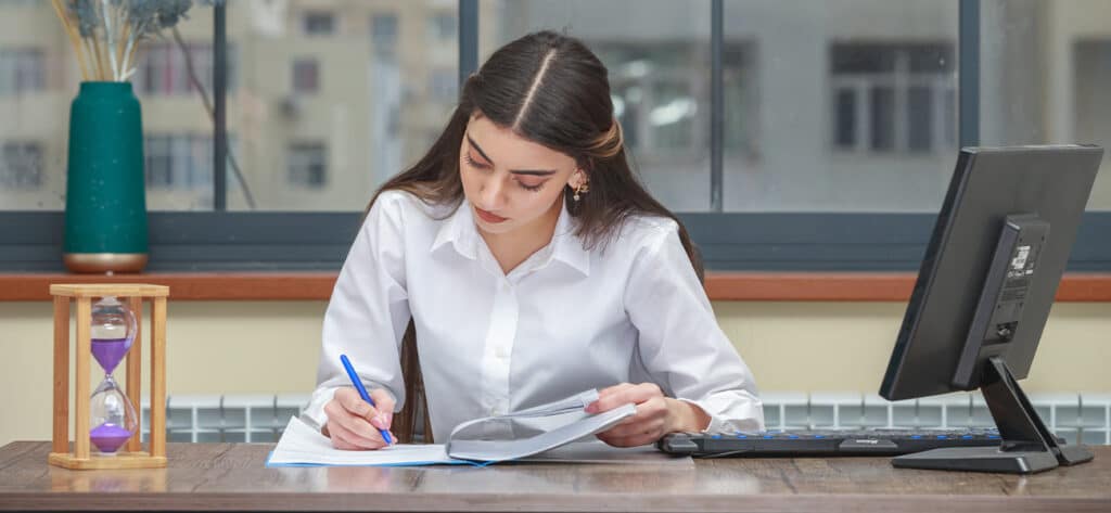Jeune femme concentrée, en chemise blanche, écrit sur son bureau près d'un sablier violet et d'un écran d'ordinateur.