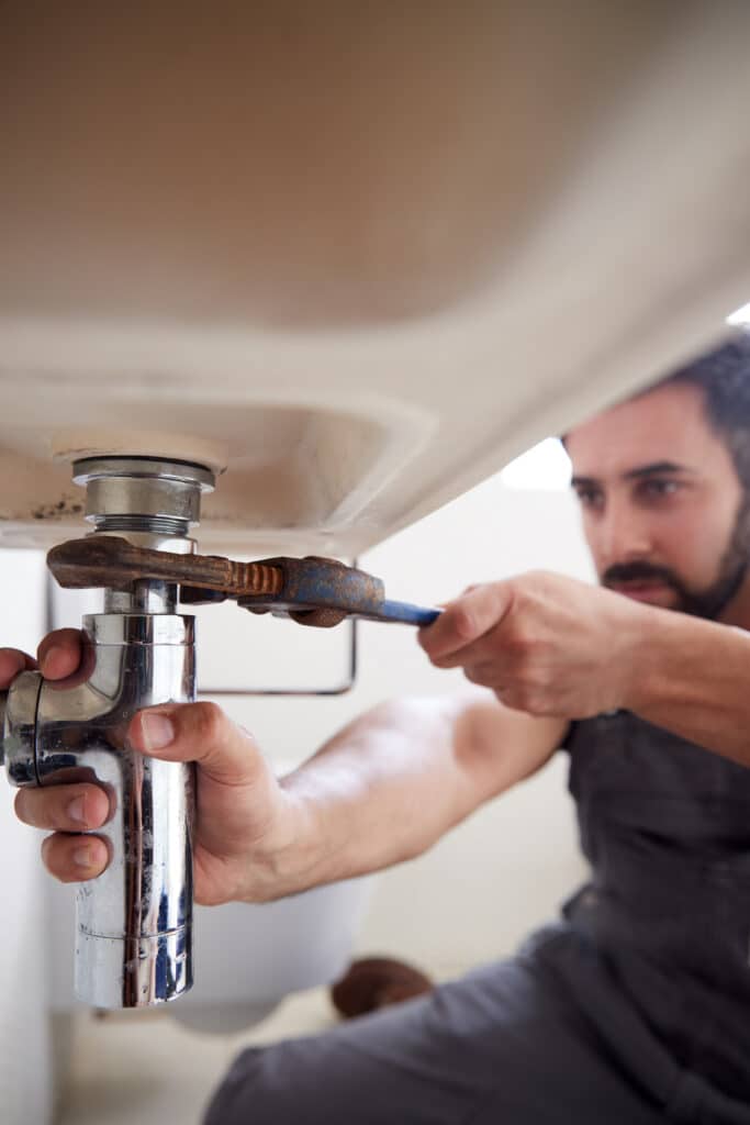 Un plombier barbu répare la tuyauterie sous un évier, utilisant une clé à molette rouillée sur un siphon chromé mouillé. Visage concentré.