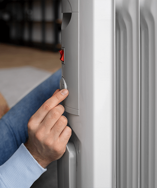 Main réglant le thermostat d'un radiateur électrique gris. Un interrupteur rouge est visible, suggérant le chauffage domestique.