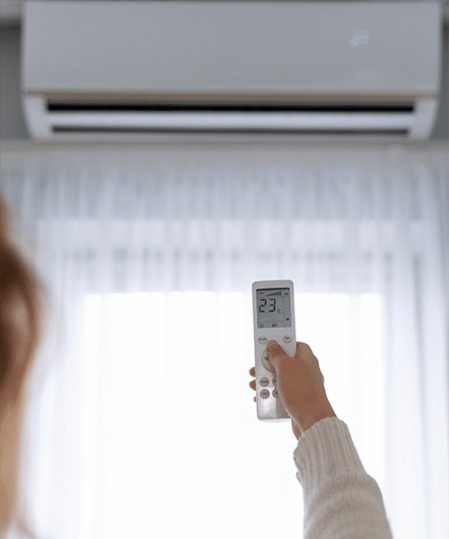 Personne utilisant une télécommande blanche pour régler la climatisation à 23°C, avec des rideaux blancs en arrière-plan.